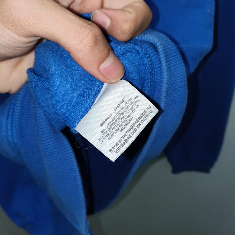 Pu&ograve; includere: Primo piano di una felpa blu reale con un'etichetta. L'etichetta contiene testo tra cui "RM856323 CA#05553" e "MADE IN VIETNAM/FABRIQUE AU VIETNAM/HECHO EN VIETNAM". La felpa &egrave; realizzata in un materiale morbido.