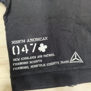 Puede incluir: Sudadera gris oscuro con texto blanco: "NORTH AMERICAN 047", "NEW ENGLAND AIR PATROL FOXBORO SCOUTS FOXBORO, NORFOLK COUNTY, MASS." Tambi&eacute;n se ve un logotipo de tri&aacute;ngulo blanco.