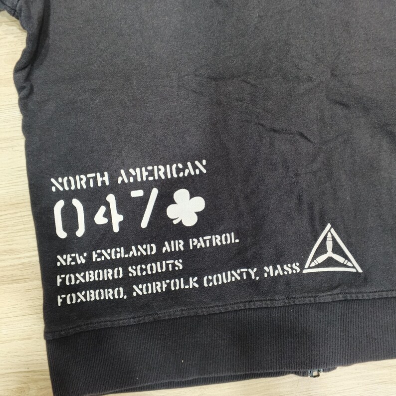 Puede incluir: Sudadera gris oscuro con texto blanco: "NORTH AMERICAN 047", "NEW ENGLAND AIR PATROL FOXBORO SCOUTS FOXBORO, NORFOLK COUNTY, MASS." Tambi&eacute;n se ve un logotipo de tri&aacute;ngulo blanco.