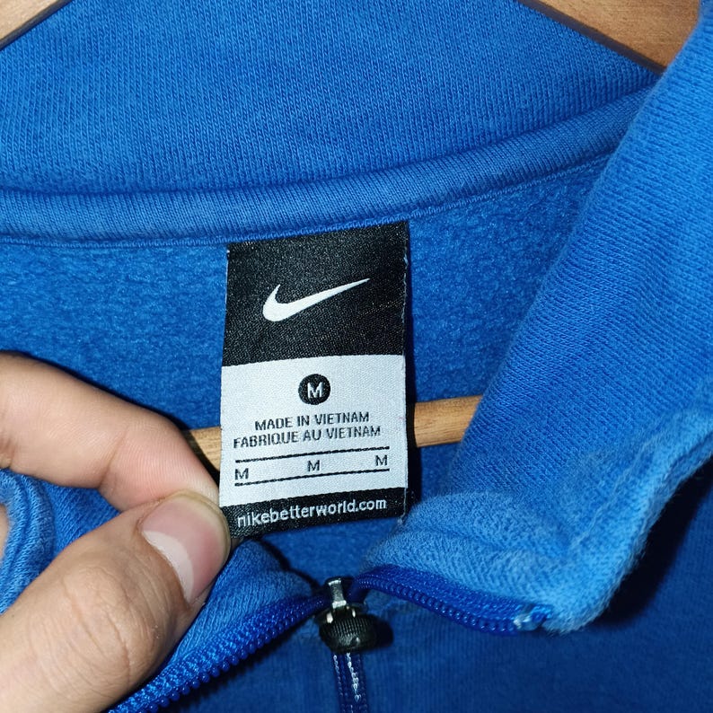 Pu&ograve; includere: Primo piano di una felpa Nike blu con logo Nike bianco e un'etichetta con la scritta "MADE IN VIETNAM FABRIQUE AU VIETNAM". L'etichetta include anche la taglia "M" e il sito web "nikebetterworld.com".