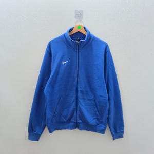Pu&ograve; includere: Giacca blu con zip e logo Nike bianco sul petto. La giacca ha un collo alto, due tasche anteriori e una cerniera a tutta lunghezza. &Egrave; appesa a una gruccia di legno.