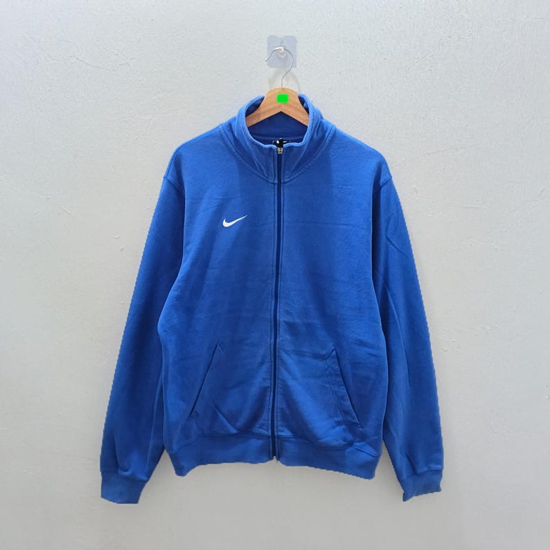 Pu&ograve; includere: Giacca blu con zip e logo Nike bianco sul petto. La giacca ha un collo alto, due tasche anteriori e una cerniera a tutta lunghezza. &Egrave; appesa a una gruccia di legno.