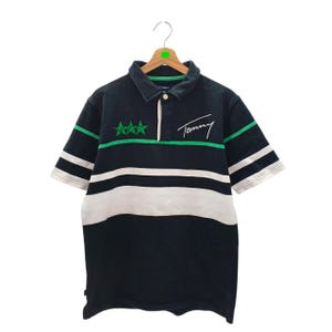 Puede incluir: Polo de color azul marino con rayas blancas y verdes y el nombre de la marca "Tommy" en escritura blanca. La camisa presenta tres estrellas verdes y un cuello blanco.
