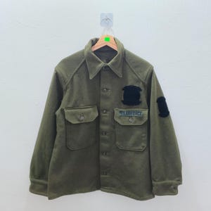 Puede incluir: Chaqueta verde oliva de estilo militar con cuello y cierre de botones. Cuenta con dos bolsillos en el pecho y parches en las mangas y el pecho. El texto "US AIR FORCE" es visible en un parche.