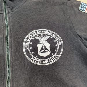 Puede incluir: Sudadera con capucha gris oscuro con cremallera y un emblema circular blanco con el texto "United States Air Force Auxiliary" y "Avirex Air Patrol". El emblema incluye un &aacute;guila y un escudo. Se ve un peque&ntilde;o parche de la bandera estadounidense.