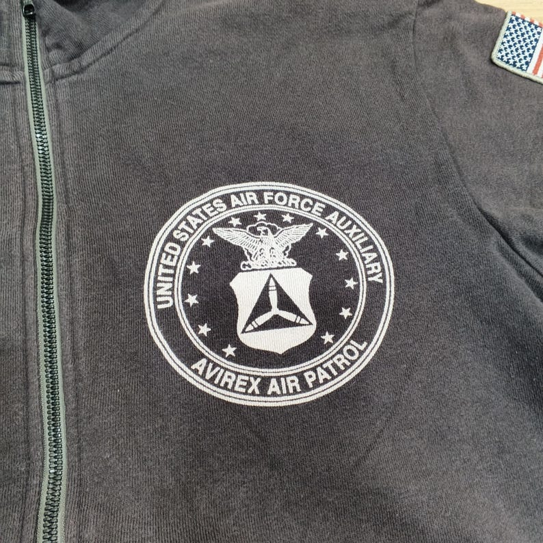 Puede incluir: Sudadera con capucha gris oscuro con cremallera y un emblema circular blanco con el texto "United States Air Force Auxiliary" y "Avirex Air Patrol". El emblema incluye un &aacute;guila y un escudo. Se ve un peque&ntilde;o parche de la bandera estadounidense.