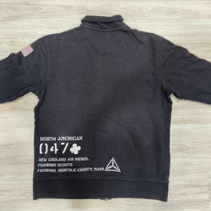 Puede incluir: Chaqueta negra con cremallera y un peque&ntilde;o parche de la bandera estadounidense en el hombro izquierdo. La parte trasera presenta texto blanco, incluyendo "NORTH AMERICAN (047)". La chaqueta tiene cuello y mangas largas.