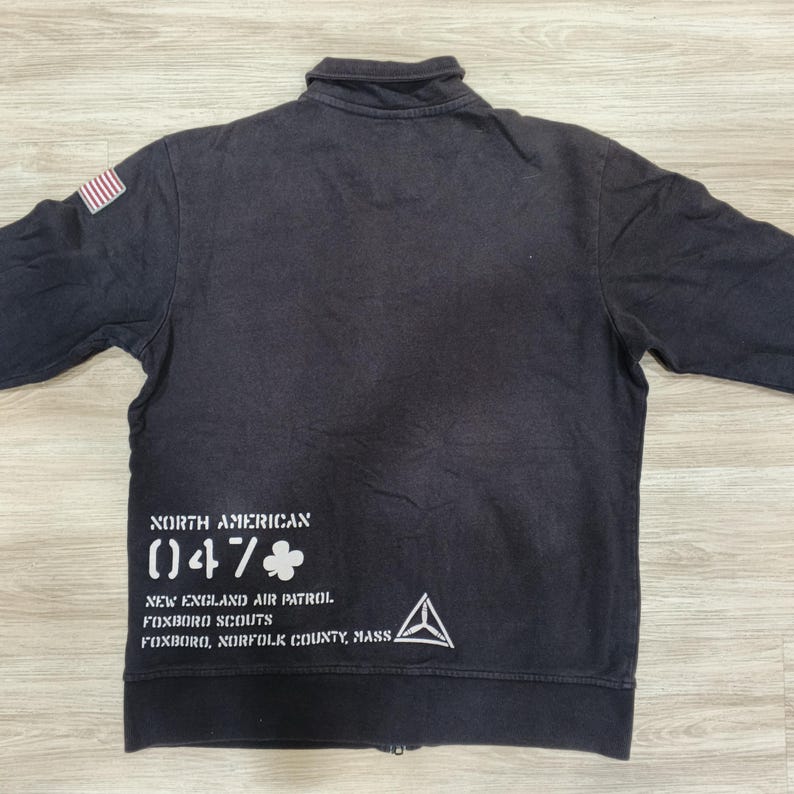 Puede incluir: Chaqueta negra con cremallera y un peque&ntilde;o parche de la bandera estadounidense en el hombro izquierdo. La parte trasera presenta texto blanco, incluyendo "NORTH AMERICAN (047)". La chaqueta tiene cuello y mangas largas.