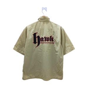 Peut inclure: Chemise beige à manches courtes avec le mot "Hawk" brodé en rouge et noir dans une police gothique au dos, avec "skateboarding" en dessous. La chemise est suspendue à un cintre blanc.