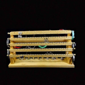 May include: Wooden jewelry organizer with four tiers of pegs for hanging earrings and a bottom shelf for storing bracelets. The organizer is made of natural wood and has a light brown finish.