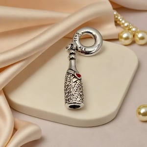 Può includere: Un charm d'argento a forma di bottiglia di champagne con un cuore rosso. La bottiglia è incastonata con piccole pietre scintillanti. Il charm ha un anello con le marcature "S925 ALE". Lo sfondo è di colore neutro.