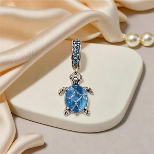 May include: A silver turtle charm with a blue, ocean-like centre. The charm has a detailed design with small blue stones on the top and around the turtle's body. The charm is displayed on a white surface with a satin fabric background.