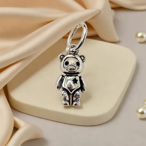 May include: A silver teddy bear charm with a loop for hanging. The bear has a star on its chest and blue eyes. The charm is displayed on a cream surface with a satin fabric in the background.