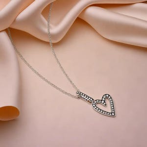 Puede incluir: Un collar de plata con un colgante en forma de corazón. El colgante está decorado con pequeñas piedras transparentes.