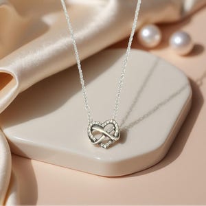 Puede incluir: Un collar plateado con un colgante en forma de corazón y un símbolo de infinito. El colgante está incrustado con pequeñas piedras preciosas transparentes. El collar se muestra sobre una superficie clara e inclinada, con un fondo neutro.