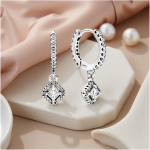 May include: Silver hoop earrings with square pendants. The hoops and pendants are adorned with small, clear gemstones. The earrings are presented on a white surface against a soft pink backdrop. The earrings are a stylish accessory.