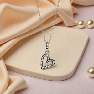 Puede incluir: Un collar plateado con un colgante en forma de corazón. El colgante está adornado con pequeños cristales transparentes, y la cadena es fina. El collar se presenta sobre una superficie clara con una tela color melocotón en el fondo.