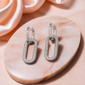 Peut inclure: Une paire de boucles d'oreilles créoles argentées avec un pendentif ovale. Les pendentifs sont recouverts de petites pierres de zircone cubique transparentes.