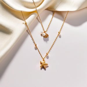 May include: A gold chain necklace with a starfish pendant and a smaller pendant with a pearl and a seashell. The necklace is on a white background.