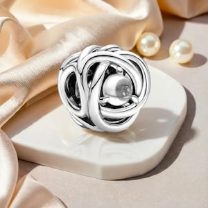 Könnte beinhalten: Ein silberner Charm-Anhänger mit einem komplizierten, ineinandergreifenden Design, das eine zentrale, transparente, kugelförmige Perle aufweist. Der Charm wird auf einer weißen, quadratischen Oberfläche vor einem weichen, neutralen Hintergrund mit zwei Perlen präsentiert.