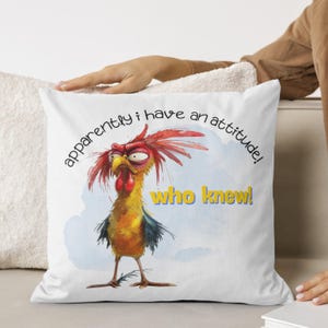 Puede incluir: Almohada blanca con un gallo de dibujos animados con cresta roja y plumas amarillas. El gallo tiene una expresi&oacute;n gru&ntilde;ona y el texto "&iexcl;aparentemente tengo una actitud! &iexcl;qui&eacute;n lo sab&iacute;a!" est&aacute; impreso en la almohada.