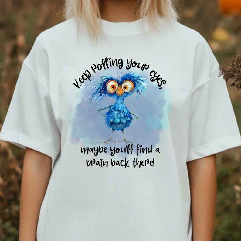 以下が含まれることがあります： 白いTシャツに、青い漫画の鳥が描かれており、"Keep rolling your eyes, maybe you'll find a brain back there!"というテキストが書かれています