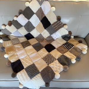 Pu&ograve; includere: Una coperta quadrata lavorata a maglia a mano con un design patchwork. La coperta presenta varie trame e motivi in tonalit&agrave; neutre di bianco, crema, grigio e marrone. &Egrave; rifinita con pompon sui bordi.