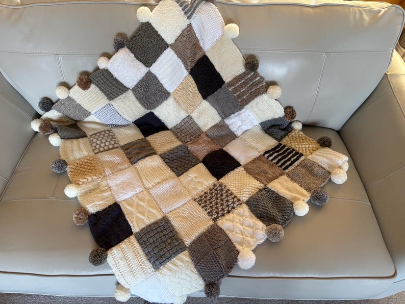 Pu&ograve; includere: Una coperta quadrata lavorata a maglia a mano con un design patchwork. La coperta presenta varie trame e motivi in tonalit&agrave; neutre di bianco, crema, grigio e marrone. &Egrave; rifinita con pompon sui bordi.