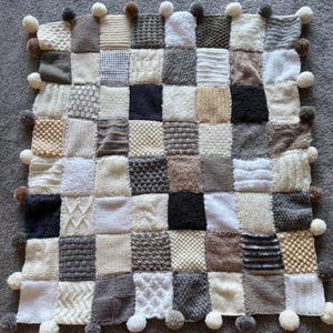 Pu&ograve; includere: Una coperta patchwork lavorata a maglia in varie tonalit&agrave; di bianco, crema, grigio e marrone. La coperta &egrave; composta da quadrati con diverse trame e motivi ed &egrave; rifinita con piccoli pompon.