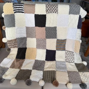 Pu&ograve; includere: Una coperta patchwork fatta a mano con una variet&agrave; di quadrati strutturati nei toni neutri di crema, grigio, marrone e nero. La coperta presenta un bordo con pompon ed &egrave; drappeggiata su una superficie.