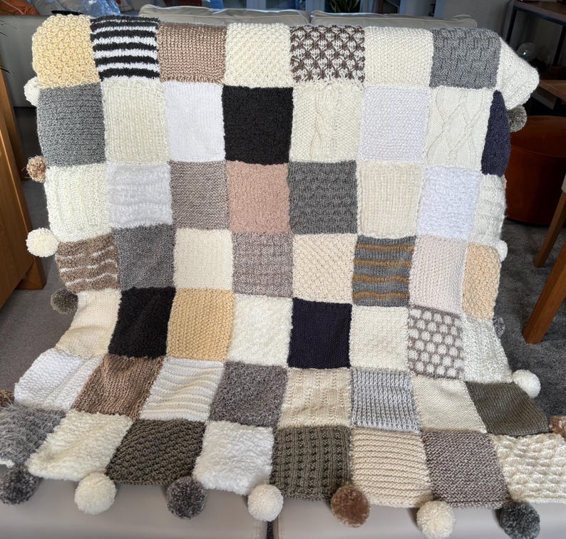 Pu&ograve; includere: Una coperta patchwork fatta a mano con una variet&agrave; di quadrati strutturati nei toni neutri di crema, grigio, marrone e nero. La coperta presenta un bordo con pompon ed &egrave; drappeggiata su una superficie.