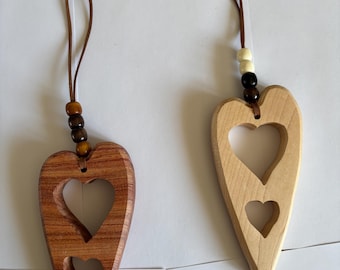 May include: Two wooden heart ornaments with a cutout design. The ornament on the left is made of dark wood with brown beads on the cord. The ornament on the right is made of light wood with white and black beads on the cord.