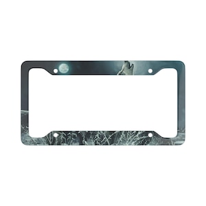 May include: A black metal licence plate frame featuring a wolf howling at the moon in a snowy forest scene. The frame has a silver finish and is designed to fit standard licence plates.