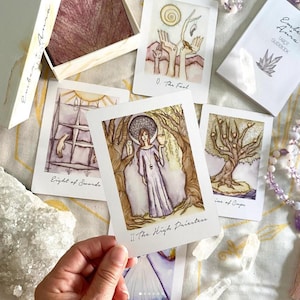 May include: A tarot card deck with watercolor illustrations. The cards feature images of The Fool, Eight of Swords, The High Priestess, and Nine of Cups. The deck is accompanied by a guidebook and a box with the words "Ember Aura" on it.