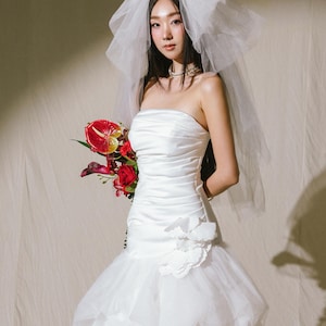 May include: A white strapless wedding dress with a ruched bodice, a layered tulle skirt, and a large white fabric flower accent at the hip. The ensemble includes a sheer white tulle veil and a bouquet of red anthurium and rose flowers. Text reads Bella.H.