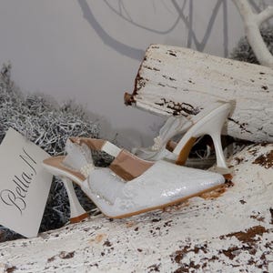 May include: White, pointed-toe slingback heels with a decorative bow and pearl accents. The heels have a unique curved design and are set on a wooden surface. The shoes are embellished with lace and rhinestones. The brand name "Bella M" is visible.