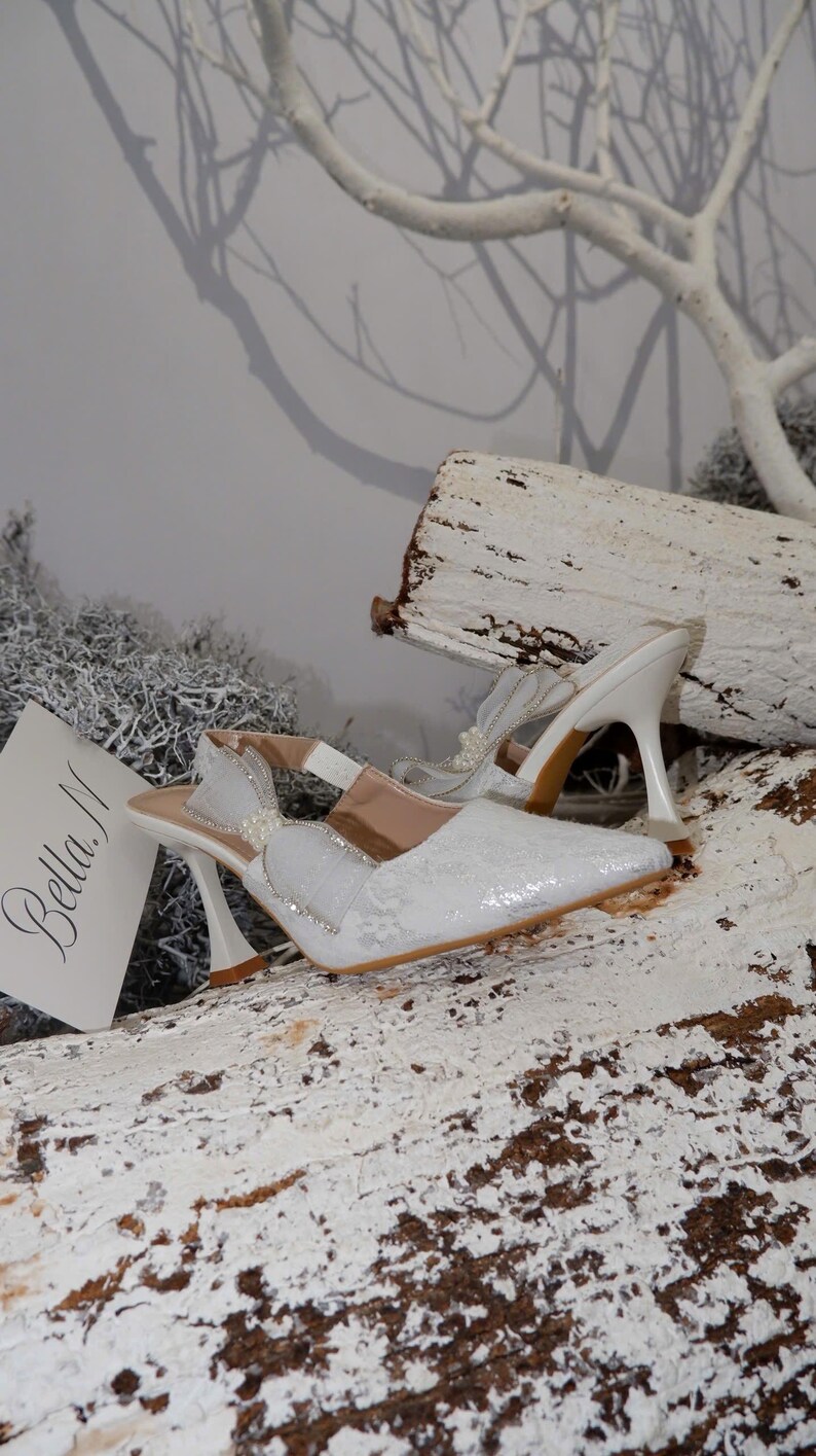 May include: White, pointed-toe slingback heels with a decorative bow and pearl accents. The heels have a unique curved design and are set on a wooden surface. The shoes are embellished with lace and rhinestones. The brand name "Bella M" is visible.