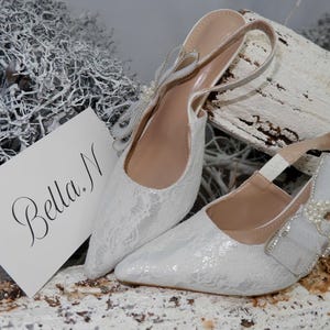 May include: A pair of white lace slingback heels with pointed toes. The shoes have a bow and pearl embellishment on the side. A card with the name "Bella N" is next to the shoes.