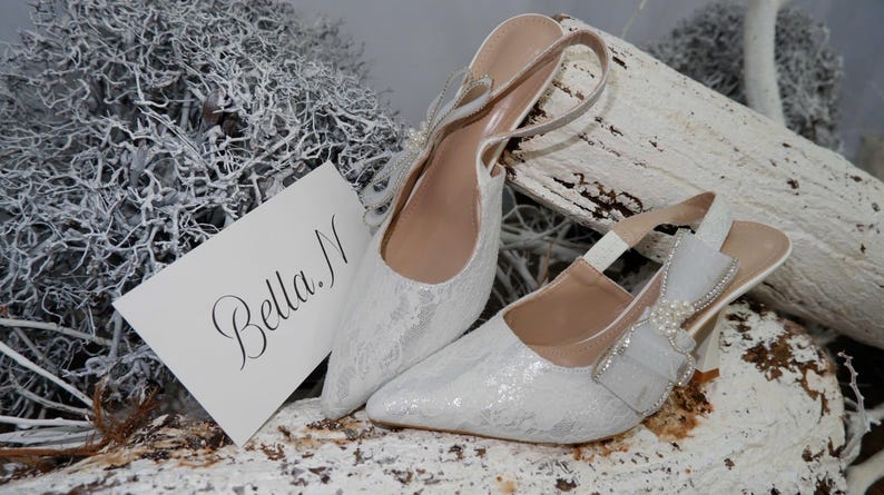 May include: A pair of white lace slingback heels with pointed toes. The shoes have a bow and pearl embellishment on the side. A card with the name "Bella N" is next to the shoes.