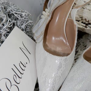May include: White lace slingback heels with a pointed toe and a decorative bow adorned with pearls. The heels have a delicate, feminine design, perfect for weddings or special occasions. A card with the name "Bella N" is visible.