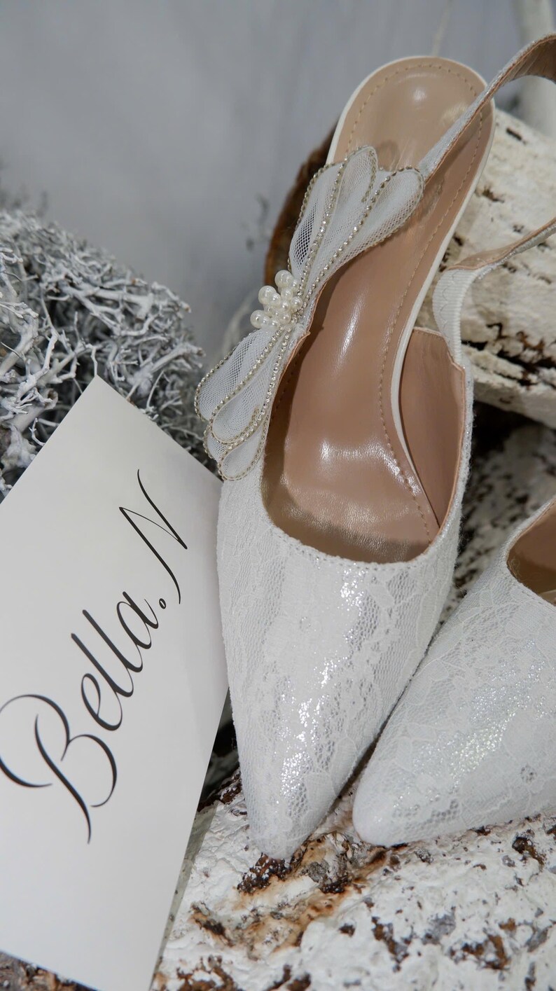 May include: White lace slingback heels with a pointed toe and a decorative bow adorned with pearls. The heels have a delicate, feminine design, perfect for weddings or special occasions. A card with the name "Bella N" is visible.