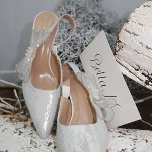May include: A pair of white, pointed-toe slingback heels with a delicate lace overlay. Each shoe features a decorative bow with pearl accents. The heels are set on a neutral background with a card that reads "Bella."