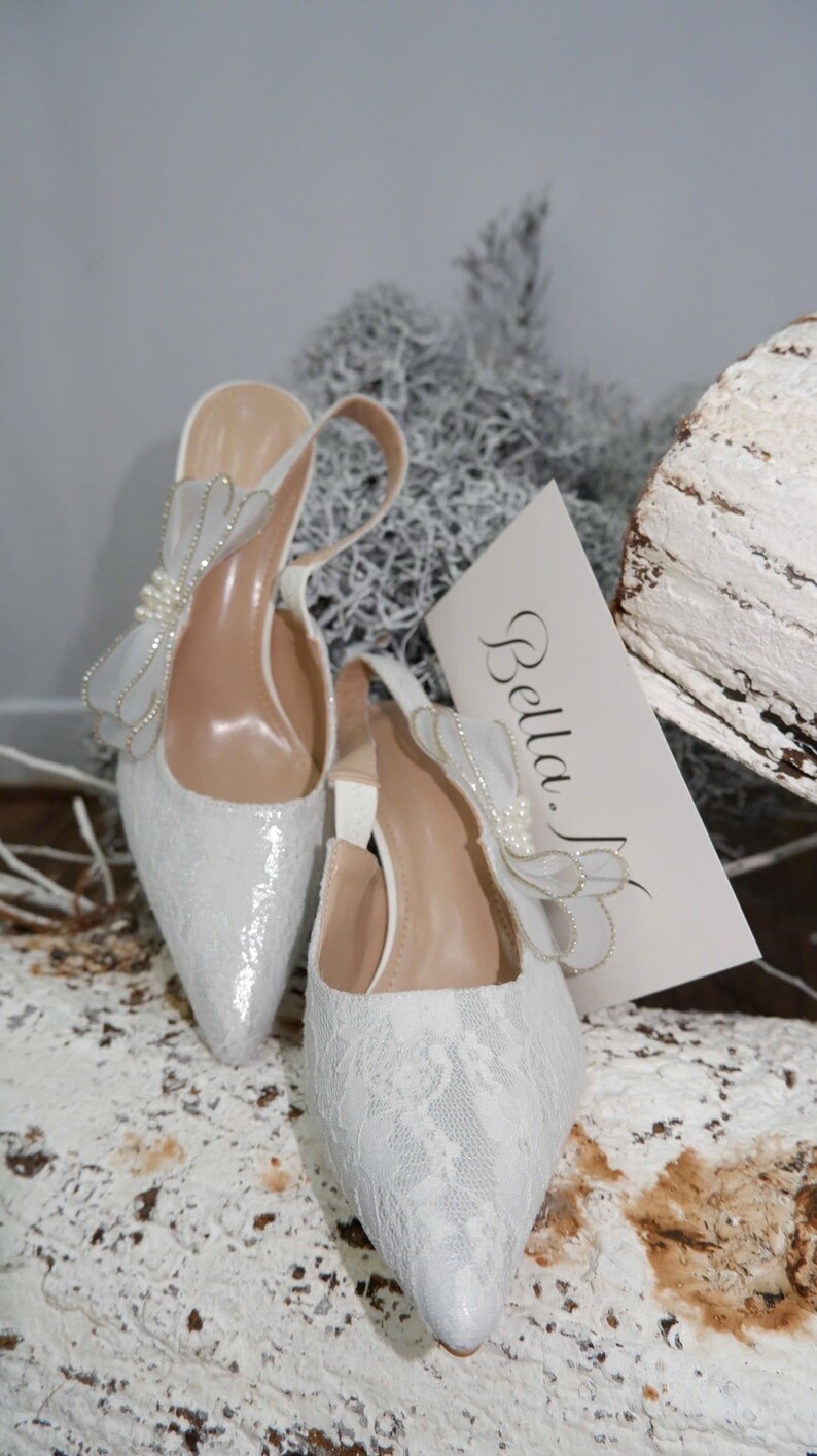 May include: A pair of white, pointed-toe slingback heels with a delicate lace overlay. Each shoe features a decorative bow with pearl accents. The heels are set on a neutral background with a card that reads "Bella."