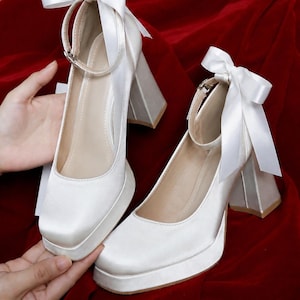 May include: A pair of white satin platform heels featuring a square toe, a thick block heel, and an ankle strap adorned with a decorative white satin ribbon bow. The shoes have a smooth finish and a light-colored interior lining.