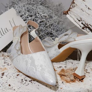 May include: White lace wedding shoes with a pointed toe and a unique heel design. The shoes feature a delicate bow detail and a strap. A card with the name "Bella" is visible.