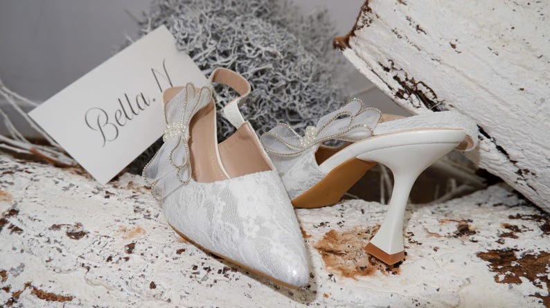 May include: White lace wedding shoes with a pointed toe and a unique heel design. The shoes feature a delicate bow detail and a strap. A card with the name "Bella" is visible.