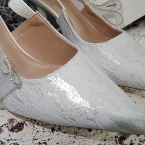 May include: A pair of elegant white lace heels with pointed toes. The shoes feature a delicate bow adorned with pearls and rhinestones. The heels are a low height, suitable for a wedding or special occasion.
