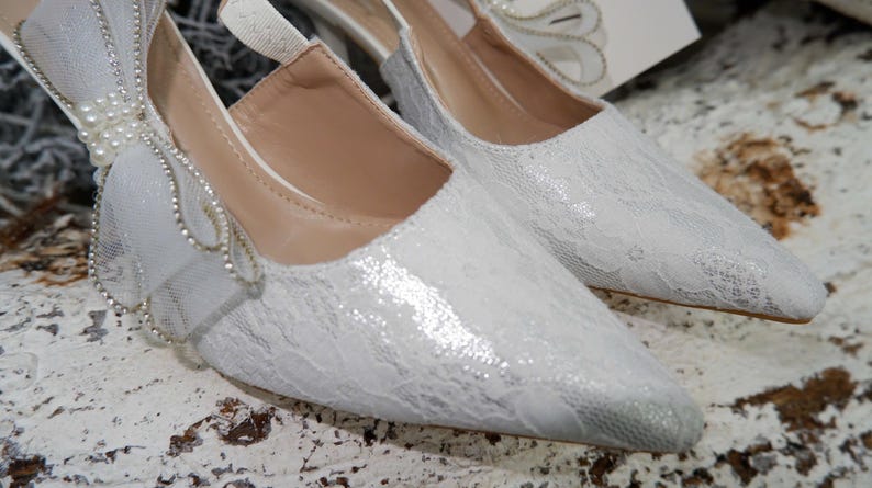 May include: A pair of elegant white lace heels with pointed toes. The shoes feature a delicate bow adorned with pearls and rhinestones. The heels are a low height, suitable for a wedding or special occasion.