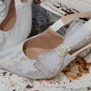 May include: White lace slingback heels with a bow and pearl embellishments. The heels have a pointed toe and a small heel. The bow is made of a sheer fabric and is adorned with rhinestones. The text "Bella N" is visible on a white card.