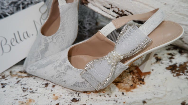 May include: White lace slingback heels with a bow and pearl embellishments. The heels have a pointed toe and a small heel. The bow is made of a sheer fabric and is adorned with rhinestones. The text "Bella N" is visible on a white card.